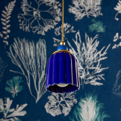 A deep, jewel-toned cobalt ceramic shade hangs like a precious object from a brass rod, its glossy surface reflecting light with an almost lacquered richness. The pleated form tapers elegantly, and the white interior creates a dramatic contrast. A cobalt sphere finial sits atop, reinforcing the jewel-box aesthetic. Against a dark navy wallpaper with delicate botanical line drawings and silvery accents, this fixture reads as pure drama—moody, luxurious, and beautifully bold.
