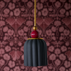A matte charcoal black ceramic shade in vertical pleats is topped with a glossy black cherry red sphere finial, separated by a brass gallery band. The two-tone composition—soft dark base with jewel-toned finial accent—creates unexpected sophistication. Against rich, damask-like wallpaper in deep mauve and burgundy tones, this fixture reads as dramatically romantic, like lighting designed for a moody, collected space.