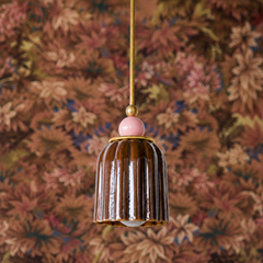 A glossy dark brown ceramic shade in vertical pleats is topped with a pale pink sphere finial, separated by brass hardware. Against a complex, multi-toned botanical wallpaper featuring dried florals, birds, and naturalistic scenes in warm browns, creams, and muted accents, this fixture reads as deeply romantic and carefully collected. The brown provides grounding; the pale pink finial adds an unexpected, gentle note of softness.