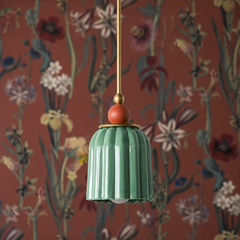 A glossy sage-green ceramic shade in vertical pleats hangs beneath a warm coral-terracotta sphere finial, trimmed in brass. Against a deep rust-toned botanical wallpaper featuring wildflowers and dried botanicals, this fixture creates a warm, naturalistic composition. The sage green and coral pairing feels effortlessly sophisticated—like colors found together in nature, pulled into an intentional design moment.