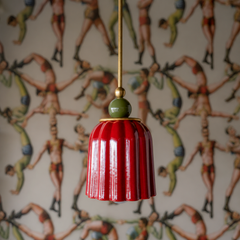 A glossy scarlet-red ceramic shade in vertical pleats is crowned with a jewel-green sphere finial, connected by brass hardware. The red is saturated and confident—reminiscent of vintage Murano glass or 1970s European ceramics. The unexpected green finial creates a sophisticated color play: jewel tones in conversation with each other. Against ornate, collected wallpaper featuring birds and botanical motifs in warm, earthy tones, this fixture reads as deliberately artistic and richly colored.
