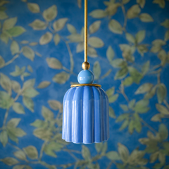 A sky-blue ceramic shade in elegant vertical pleats hangs like a gentle beacon from a brass rod, its glossy glaze catching light in soft, luminous tones. The matching pale blue sphere finial sits atop, creating a monochromatic composition that feels serene rather than monotonous. Against botanical wallpaper with soft greens and delicate botanical line drawings, this fixture reads as dreamy and collected—the kind of lighting you find in spaces designed with genuine thoughtfulness.