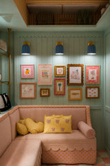 A cheerful kids playroom reading nook with mint beadboard walls and scalloped trim, an orange-and-white striped ceiling, and a playful gallery wall. Three unglazed blue ceramic Marcella wall sconces in a soft medium pastel blue add warm light above a blush pink sofa, giving the space a preppy, grandmillennial-meets-vintage feel that works beautifully in a nursery, kids bedroom, or family hangout nook.