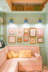 A cheerful kids playroom reading nook with mint beadboard walls and scalloped trim, an orange-and-white striped ceiling, and a playful gallery wall. Three unglazed blue ceramic Marcella wall sconces in a soft medium pastel blue add warm light above a blush pink sofa, giving the space a preppy, grandmillennial-meets-vintage feel that works beautifully in a nursery, kids bedroom, or family hangout nook.