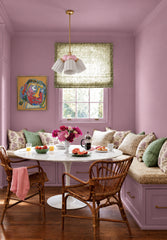 A breakfast nook painted head to toe in dusty lilac, from the walls and ceiling to the trim and built-in banquette, so the whole corner feels wrapped in color. Block-print pillows in lavender, olive and tan, rattan armchairs, a bold abstract portrait and a bowl of peonies keep it collected and a little feminine. A small chandelier with white fluted ceramic shades, pink glass globes and a brass stem hangs low over the tulip table, soft against all that lilac.