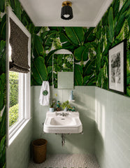Maximalist tropical bathroom with oversized green banana leaf wallpaper, pale green square tile, black and white mosaic floor, a white wall-mounted sink, arched mirror, and woven accents. The unglazed black Marcella Flushmount with brass canopy adds a small but graphic ceiling light to the room, with a ribbed ceramic shade, scalloped edge, and visible white globe bulb. A strong fit for bathroom ceiling lighting, powder room lighting, jungle wallpaper interiors, black and brass flush mounts, ceramic ceiling 