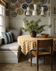Cozy breakfast nook with a built-in banquette, round table, and mustard gingham tablecloth, styled with a ceramic bowl centerpiece and a large leafy arrangement. A curated gallery wall of vintage plates and blue-and-white ceramics adds collected, traditional charm above white beadboard wainscoting. The Sazerac Stitches Margot Chain Chandelier in cream with warm brass chain links brings soft, sculptural statement lighting for a cottage kitchen, French country, coastal grandma, and vintage-inspired dining cor