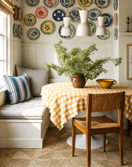 Sunlit dining nook styled with a classic plate wall, white paneled wainscoting, and a comfy banquette bench with striped pillows. The round breakfast table is draped in a mustard checkered tablecloth for an easy cottagecore, farmhouse, and traditional kitchen look. Overhead, a cream and brass chain-hung chandelier with three tapered shades creates a refined, warm glow and adds designer lighting style for a breakfast nook, kitchen banquette, or small dining space.