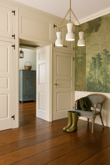 The Margot chandelier, featuring four unglazed ceramic hourglass shades and brass chain detailing, hangs elegantly in a transitional entryway with floor-to-ceiling cream paneled cabinetry, a scenic green botanical mural, warm wood floors, and vintage-inspired accents like a wicker chair, olive green wellies, and a slate blue cabinet in the adjoining room—blending modern lighting design with timeless, cottagecore interiors.