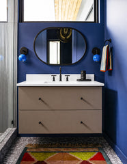 Color-drenched modern bathroom with deep blue walls, a floating taupe vanity, black oval mirror, matte black faucet, terrazzo-style flooring, and a colorful geometric bath rug. A pair of Mediterranean Blue and Black Loa sconces flank the mirror, adding glossy blue wall lighting and playful contrast to the sleek bathroom vanity. This image works for modern blue bathroom ideas, colorful bathroom lighting, contemporary vanity sconces, dopamine decor, kids bathroom design, and bold powder room inspiration.