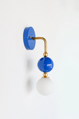The Ida Sconce pairs a brass hooked arm with a glossy Mediterranean blue glass ball and a soft white opal glass globe at the bulb, giving it a sculptural, almost jewelry-like presence on the wall. It works well as a bedside light in place of a table lamp, beside a bathroom mirror, or anywhere a single statement sconce makes more sense than a matching pair.