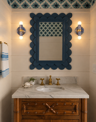 This powder room shows two blue hand-marbled Thalia sconces glowing on either side of a deep blue scalloped mirror. With warm brass accents, a marble countertop, wood vanity, and graphic blue wallpaper overhead, the space has a fresh coastal vibe with a slightly eclectic twist. The lit sconces highlight the soft ambient glow they bring to a bathroom or powder room.