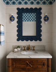 A pair of blue and gold marbled Thalia sconces flank a bold blue scalloped mirror in a playful powder room. The space features creamy wall paneling, a marble vanity top, warm brass faucet hardware, and a blue star-patterned wallpaper on the ceiling and mirror inset. The rich blue marbling gives these handmade sconces a coastal, ocean-inspired feel while still fitting eclectic and maximalist interiors.