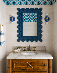A pair of blue and gold marbled Thalia sconces flank a bold blue scalloped mirror in a playful powder room. The space features creamy wall paneling, a marble vanity top, warm brass faucet hardware, and a blue star-patterned wallpaper on the ceiling and mirror inset. The rich blue marbling gives these handmade sconces a coastal, ocean-inspired feel while still fitting eclectic and maximalist interiors.