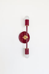 Medium Thalia Sconce shown vertically in a glossy Black Cherry finish with warm brass hardware and two white globe bulbs. This colorful two-light wall sconce has a slim, modern silhouette that works beautifully as bathroom vanity lighting, powder room lighting, hallway lighting, or a bold accent in colorful interiors with vintage, midcentury, Art Deco, or grandmillennial style.