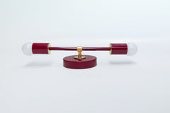 Medium Thalia Sconce shown horizontally with a glossy Black Cherry finish, brass hardware, and white globe bulbs. This two-light wall sconce has a clean, linear shape that works well above bathroom mirrors, between double vanities, or in colorful powder rooms. The deep burgundy-red finish adds a dramatic pop of color while the brass details give it a warm vintage-modern feel.