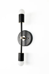 Vertical Medium Thalia wall sconce in matte black with polished chrome hardware, featuring a slim center rod, round black backplate, and two exposed frosted globe bulbs on top and bottom, modern minimal double light wall fixture for bathroom vanity lighting, hallway sconces, bedside reading lights, and contemporary mid century modern interiors