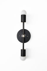 Front view of the Medium Thalia Sconce in matte black with chrome hardware, a minimalist modern double bulb wall sconce with two exposed globe bulbs, round backplate, and vertical bar, ideal as bathroom sconces flanking a mirror, hallway lighting, or bedroom bedside wall lights in modern, mid century, and high-contrast interiors.