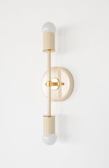Angled side view of the Medium Thalia sconce in cream with brass accents, a vertical double bulb wall sconce with exposed globe bulbs, slim stem, and round backplate, warm modern lighting style that fits mid century modern, contemporary, and elevated eclectic interiors