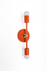 Side-angle product photo of the Medium Thalia Sconce mounted vertically in Flame orange with polished chrome accents and white globe bulbs at the top and bottom. This colorful wall sconce has a clean, geometric shape that feels modern, mid century, and slightly industrial, making it a fun choice for bathroom mirrors, powder rooms, bedside lighting, or eclectic hallways.