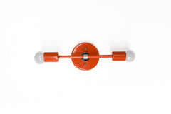 Medium Thalia Sconce shown horizontally on a white wall in a glossy Flame orange finish with chrome hardware and two exposed white globe bulbs. The slim bar shape, round backplate, and bright red-orange color give this two-light wall sconce a playful modern look for bathroom vanity lighting, kids bathrooms, powder rooms, hallways, or colorful retro interiors.