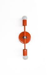 Front view of the Medium Thalia Sconce in a bold Flame orange finish with chrome hardware, a round backplate, straight center rod, and two white globe bulbs. The narrow vertical design works well beside a bathroom mirror, between double vanities, in a hallway, or as a colorful bedroom wall light for spaces that need a bright pop of warm red-orange color.