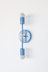 Studio photo of the Medium Thalia Sconce in soft Hydrangea blue with chrome hardware and white globe bulbs. This colorful two-light wall sconce has a slim vertical shape that works beautifully for bathroom vanity lighting, kids bathrooms, powder rooms, hallways, and playful modern interiors.