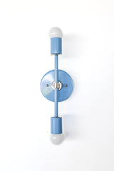 Front view of a Hydrangea blue Medium Thalia Sconce with polished chrome details, shown on a clean white wall. The narrow vertical silhouette and double globe bulbs make it a great colorful wall light for bathroom mirrors, powder rooms, kids spaces, hallway lighting, and cheerful custom interiors.