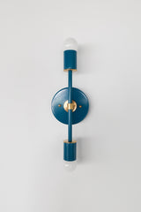 Front-facing studio photo of the Medium Thalia Sconce in a deep Lagoon blue finish with brass hardware and two white globe bulbs. This colorful vertical wall sconce has a slim, modern shape with a round backplate, making it a strong choice for bathroom vanity lighting, powder rooms, kids bathrooms, hallways, bedrooms, and interiors that need a polished pop of teal blue.