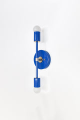Mediterranean Blue and chrome Medium Thalia Sconce shown from an angled side view on a clean white wall. This slim vertical two-light wall sconce features a glossy cobalt blue powder-coated finish, polished chrome hardware, a round backplate, and exposed white globe bulbs at the top and bottom. A colorful modern bathroom vanity light for kids bathrooms, powder rooms, hallways, bedrooms, coastal interiors, eclectic spaces, and playful blue-and-white decor.