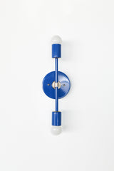 Straight-on studio photo of the Mediterranean Blue Medium Thalia Sconce with chrome hardware and two exposed globe bulbs. This vertical wall sconce has a simple modern shape with a glossy blue powder-coated finish, making it a fun choice for blue bathroom lighting, colorful vanity sconces, kids bathroom decor, coastal powder rooms, modern hallways, and playful contemporary interiors.