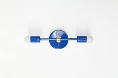 Straight-on studio photo of the Mediterranean Blue Medium Thalia Sconce with chrome hardware and two exposed globe bulbs. This horizontal wall sconce has a simple modern shape with a glossy blue powder-coated finish, making it a fun choice for blue bathroom lighting, colorful vanity sconces, kids bathroom decor, coastal powder rooms, modern hallways, and playful contemporary interiors.