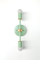 Front-facing Medium Thalia wall sconce in mint with warm brass hardware, designed with a vertical bar, circular backplate, and dual globe bulbs. The soft mint enamel and brass details bring a vintage-modern look suited to mid century modern interiors, pastel maximalist decor, bathroom vanity wall lighting, dining nook accent lighting, bedroom sconces, and boutique hotel style interiors.