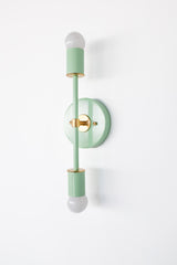 Three-quarter side view of the mint Medium Thalia sconce with brass accents, showing the depth of the round backplate and slim vertical frame with top and bottom globe bulbs. This handmade style statement wall light works as a colorful accent for powder room vanity lighting, hallway wall lighting, nursery or kids room decor, artful modern interiors, and eclectic maximalist spaces.