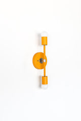 Side view of the Medium Thalia Sconce in a cheerful mustard yellow finish with chrome hardware and two white globe bulbs. This slim two-light wall sconce has a modern, playful shape that works beautifully in colorful bathrooms, kids bathrooms, powder rooms, hallways, and eclectic interiors with midcentury, retro, or Art Deco-inspired style.