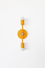 Front view of the Medium Thalia Sconce in a bold mustard yellow powder-coated finish with polished chrome hardware and two globe bulbs. The clean vertical silhouette makes it a great colorful bathroom vanity sconce, hallway light, or powder room wall fixture for spaces that need a warm pop of yellow with a modern vintage feel.