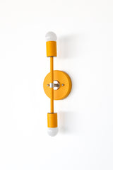 Angled view of the Medium Thalia Sconce in mustard yellow with chrome hardware, shown mounted vertically on a simple white wall. This colorful two-light wall fixture brings a happy retro accent to bathrooms, powder rooms, kids spaces, hallways, and maximalist or midcentury-inspired interiors while still feeling clean and streamlined.