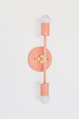 Angled studio product photo of the Medium Thalia Sconce in Peach with brass hardware and two white globe bulbs. This view highlights the fixture’s projection from the wall, round backplate, brass center mount, brass accent rings, and glossy peach powder-coated finish.