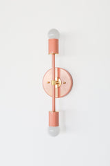 Side-angle product photo of the Medium Thalia Sconce in glossy Peach with brass hardware, shown mounted vertically against a simple white wall. The image emphasizes the fixture’s slim linear shape, round wall plate, two cylindrical socket covers, white globe bulbs, and warm brass details.