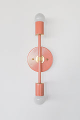Front-facing product photo of the Medium Thalia Sconce in glossy Peach with warm brass hardware, mounted vertically on a clean white wall. The image shows the round backplate, slim center rod, cylindrical socket covers, two white globe bulbs, and soft coral-peach powder-coated finish.