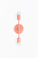 Front Facing product photo of the Medium Thalia Sconce in glossy Peach with chrome hardware and two white globe bulbs. The sconce is mounted vertically on a clean white wall, showing the round backplate, slim center rod, cylindrical socket covers, reflective chrome details, and soft coral-pink powder-coated finish.