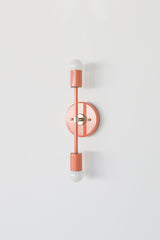 Side-angle product photo of the Medium Thalia Sconce in glossy Peach with chrome hardware and two white globe bulbs. The clean white background keeps the focus on the fixture’s projection, round backplate, vertical silhouette, cylindrical socket covers, and playful coral-toned powder-coated finish.