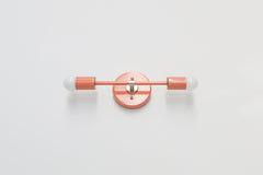 Studio product photo of the Medium Thalia Sconce in Peach with chrome hardware, shown mounted horizontally against a simple white wall. This view highlights the fixture’s flippable design, round wall plate, long linear arm, two globe bulbs, glossy peach finish, and polished chrome center mount.
