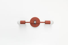 Medium Thalia Sconce shown mounted horizontally on a clean white wall in a warm terra cotta finish with polished chrome accents and two exposed white globe bulbs. The slim bar shape, round backplate, and simple cylinder sockets give this colorful wall light a modern, slightly retro look that works well as bathroom vanity lighting, over-mirror lighting, hallway lighting, or a playful wall sconce for kids rooms, powder rooms, and colorful interiors.