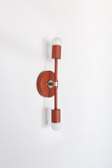 Medium Thalia Sconce shown vertically in a terra cotta finish with chrome hardware and two white globe bulbs. The long, narrow silhouette makes it easy to use beside a mirror, over a nightstand, along a hallway, or in a small bathroom where a colorful wall light needs to feel slim and intentional. The warm red-orange color adds an earthy pop against white walls and pairs well with marble, chrome, wood tones, checkerboard tile, and modern traditional interiors.