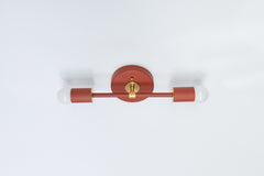 Side-angle product photo of the Terra Cotta and brass Medium Thalia Sconce mounted vertically on a white wall. The image highlights the fixture’s depth, round backplate, straight center rod, cylindrical bulb holders, brass bracket, and soft white globe bulbs. This colorful wall sconce is a strong fit for bathroom mirror lighting, powder room sconces, bedside wall lights, hallway accent lighting, and interiors that mix warm earthy color with classic brass details.