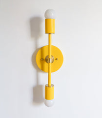 Medium Thalia Sconce in glossy yellow with brass center detailing, shown front-facing as a vertical double-light wall sconce with two white globe bulbs. Colorful modern wall lighting with a clean geometric silhouette, ideal for bathroom vanity lighting, hallway sconces, bedroom wall lights, and mid century modern or maximalist interiors.