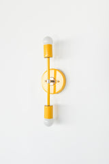Medium Thalia Sconce in bright yellow with polished chrome accents, photographed straight-on against a light neutral wall. This vertical two-light wall fixture features exposed globe bulbs and a slim architectural profile, perfect for modern bathroom sconces, contemporary hallway lighting, colorful bedroom wall lights, and playful eclectic home decor.
