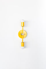 Medium Thalia Sconce in bright yellow with polished chrome accents, photographed straight-on against a light neutral wall. This vertical two-light wall fixture features exposed globe bulbs and a slim architectural profile, perfect for modern bathroom sconces, contemporary hallway lighting, colorful bedroom wall lights, and playful eclectic home decor.