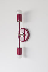 Medium Thalia Sconce shown vertically from the side with a glossy Black Cherry finish, chrome hardware, and two white globe bulbs. This slim two-light wall sconce brings a colorful modern look to bathrooms, powder rooms, hallways, bedrooms, and vanity areas, with a deep red-burgundy color that pairs beautifully with marble, white walls, patterned wallpaper, and polished chrome fixtures.